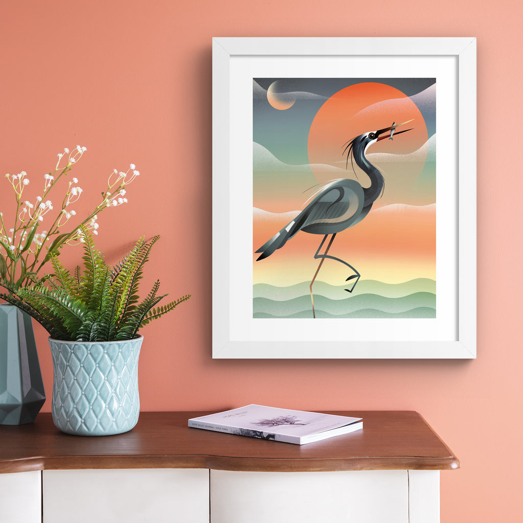 Striking art print featuring a grey heron posing in front of a stunning red sunset by the water. Art print is hung up on a pink wall.