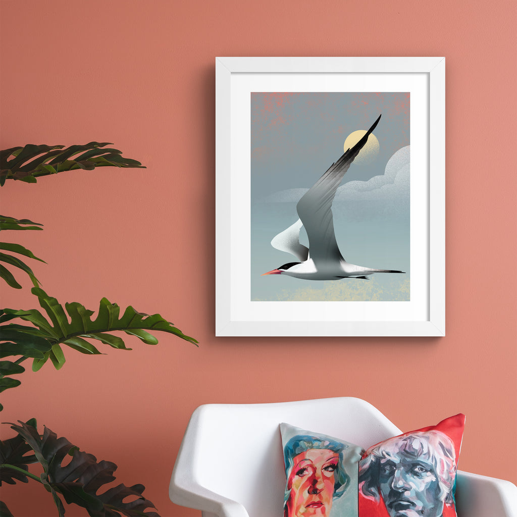 Striking art print featuring a grey tern flying in front of a moody blue sky. Art print is hung up on a pink wall.