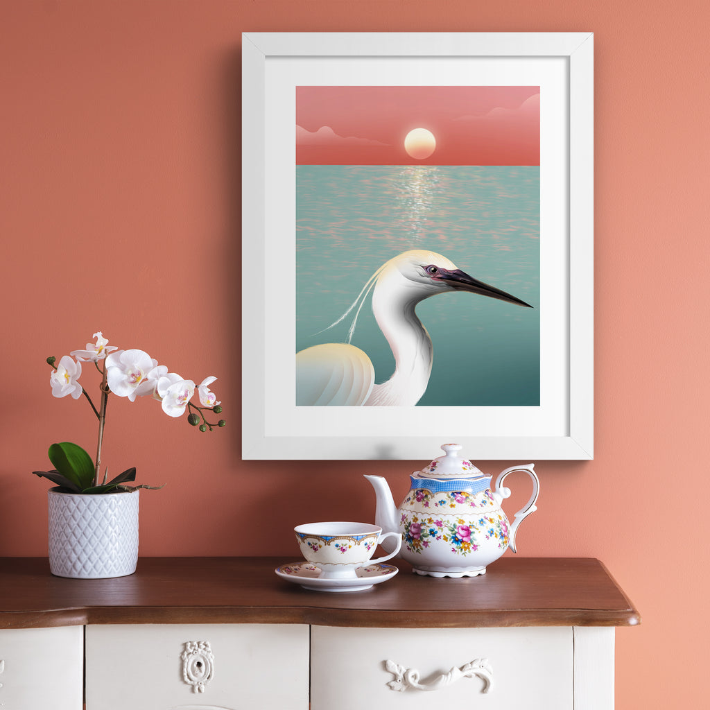 Striking art print featuring a white egret posing in from of the ocean, as the sun sets on the horizon. Art print is hung up on a pink wall.