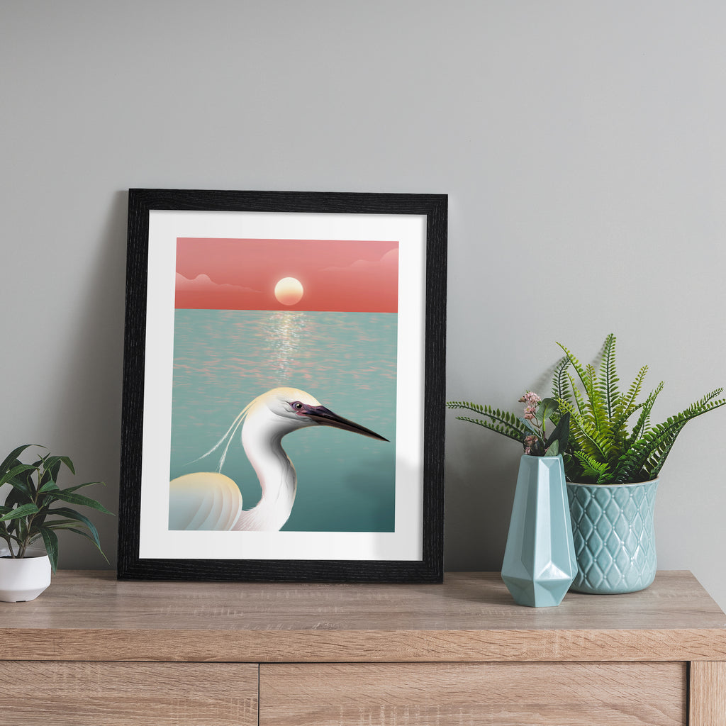 Striking art print featuring a white egret posing in from of the ocean, as the sun sets on the horizon. Art print is leaning against a grey wall.