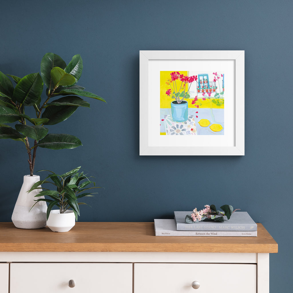 Vibrant art print featuring a vase of pink flowers next to yellow fruit and walls. Art print is hung up on a deep blue wall.