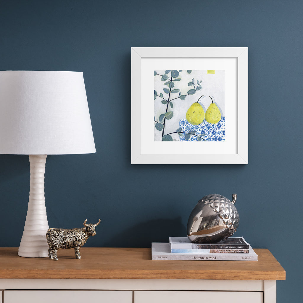 Minimalistic art print featuring two pears laid out on a blue patterned tablecloth. Art print is hung up on a dark blue wall.