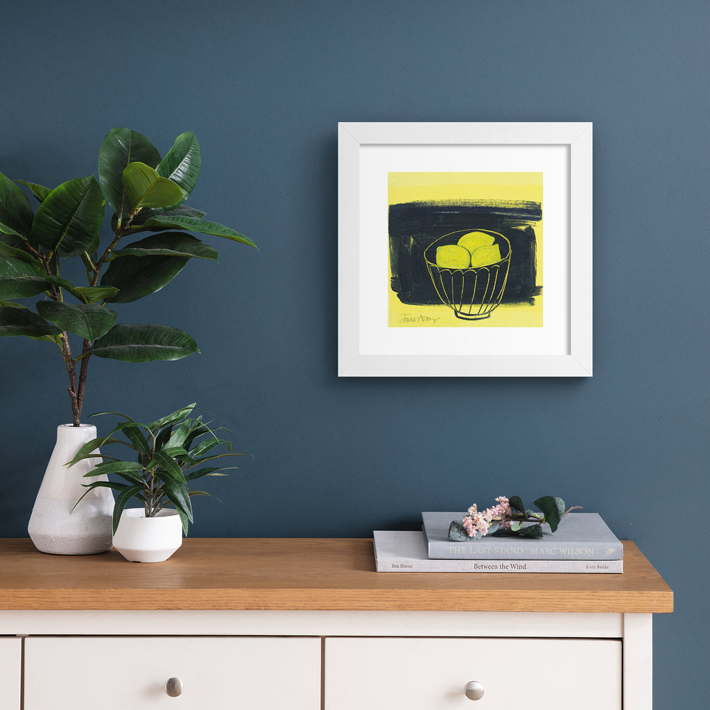 Minimalistic art print featuring lemons in a fruit bowl in front of a bright yellow and black background. Art print is hung up on a dark blue wall.