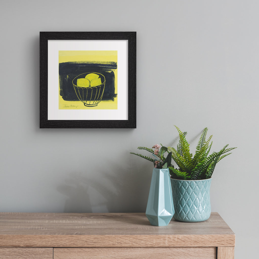 Minimalistic art print featuring lemons in a fruit bowl in front of a bright yellow and black background. Art print is hung up on a grey wall.