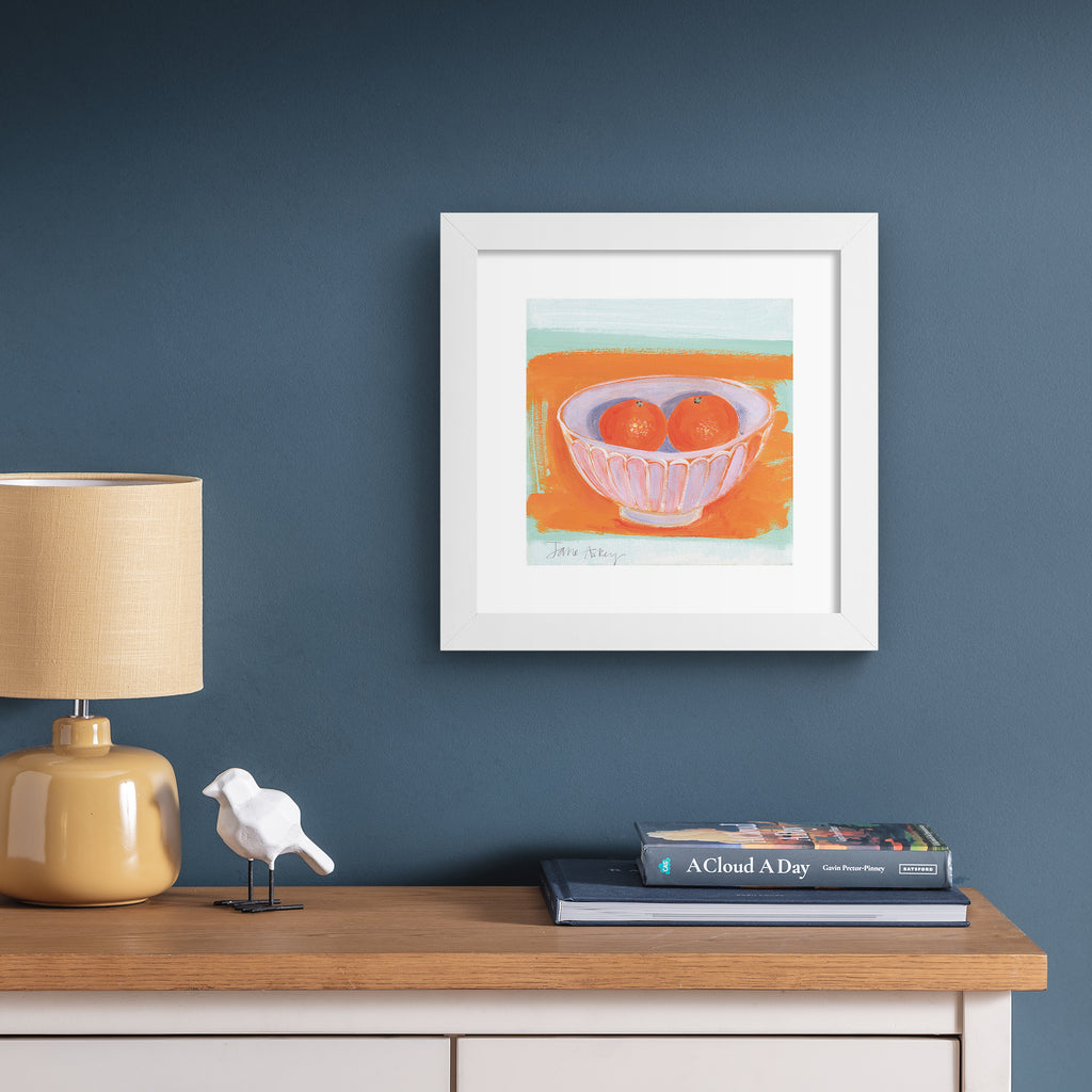 Minimalistic art print featuring oranges in a fruit bowl in front of a bright orange and blue background. Art print is hung up on a blue wall.