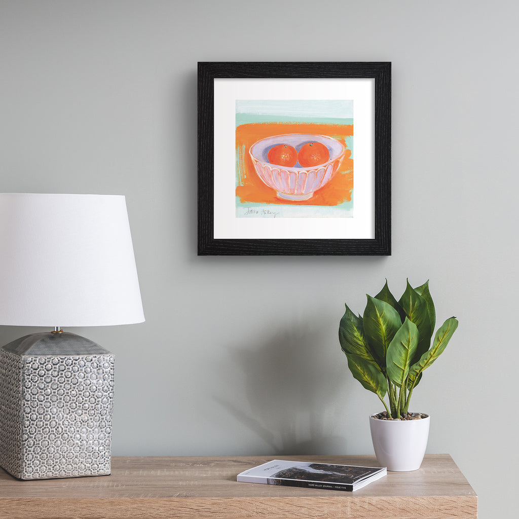Minimalistic art print featuring oranges in a fruit bowl in front of a bright orange and blue background. Art print is hung up on a beige wall.