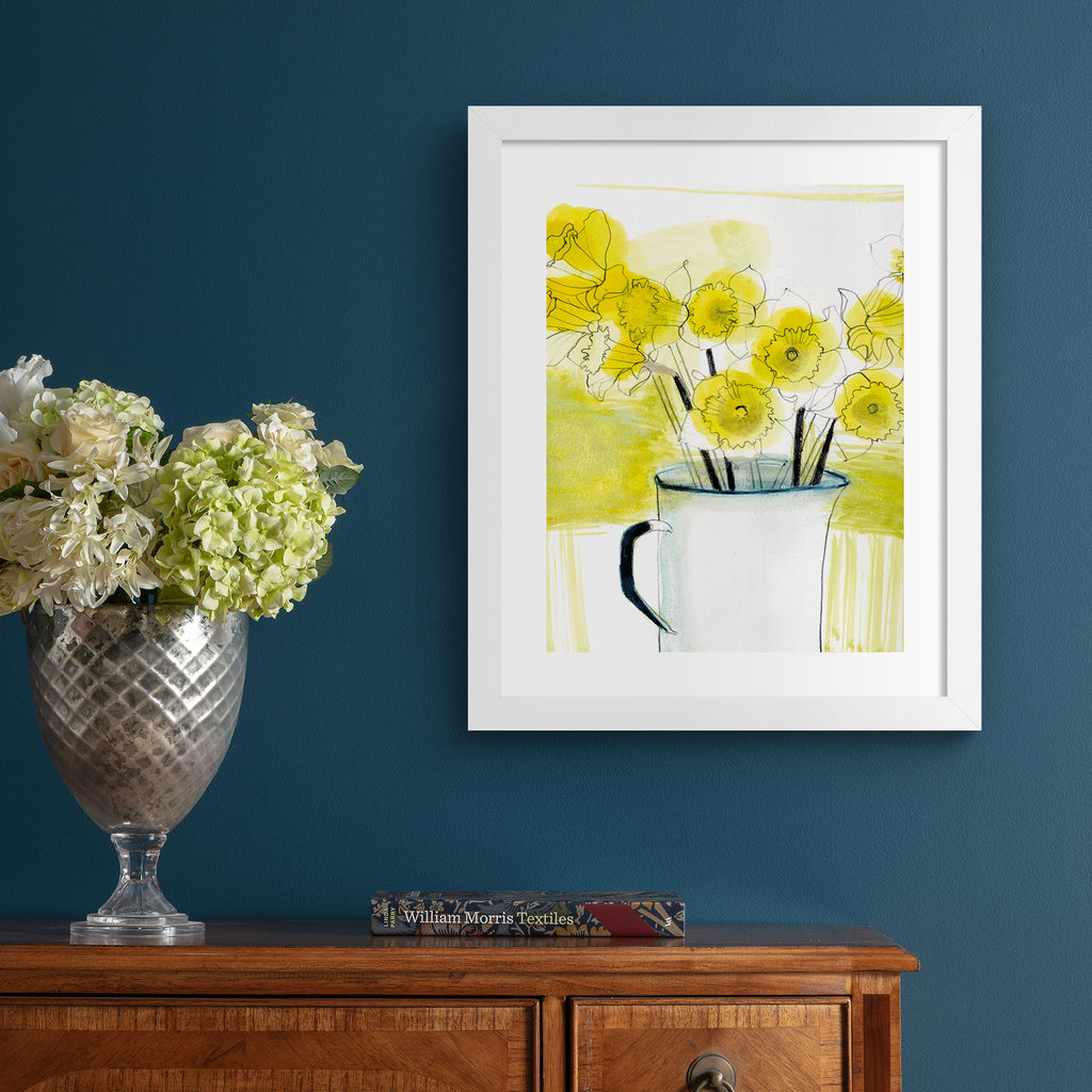 Beautiful yellow art print illustration featuring daffodils spilling out of an enamel jug, with a blended yellow and white background. Art print is hung up on a blue wall.