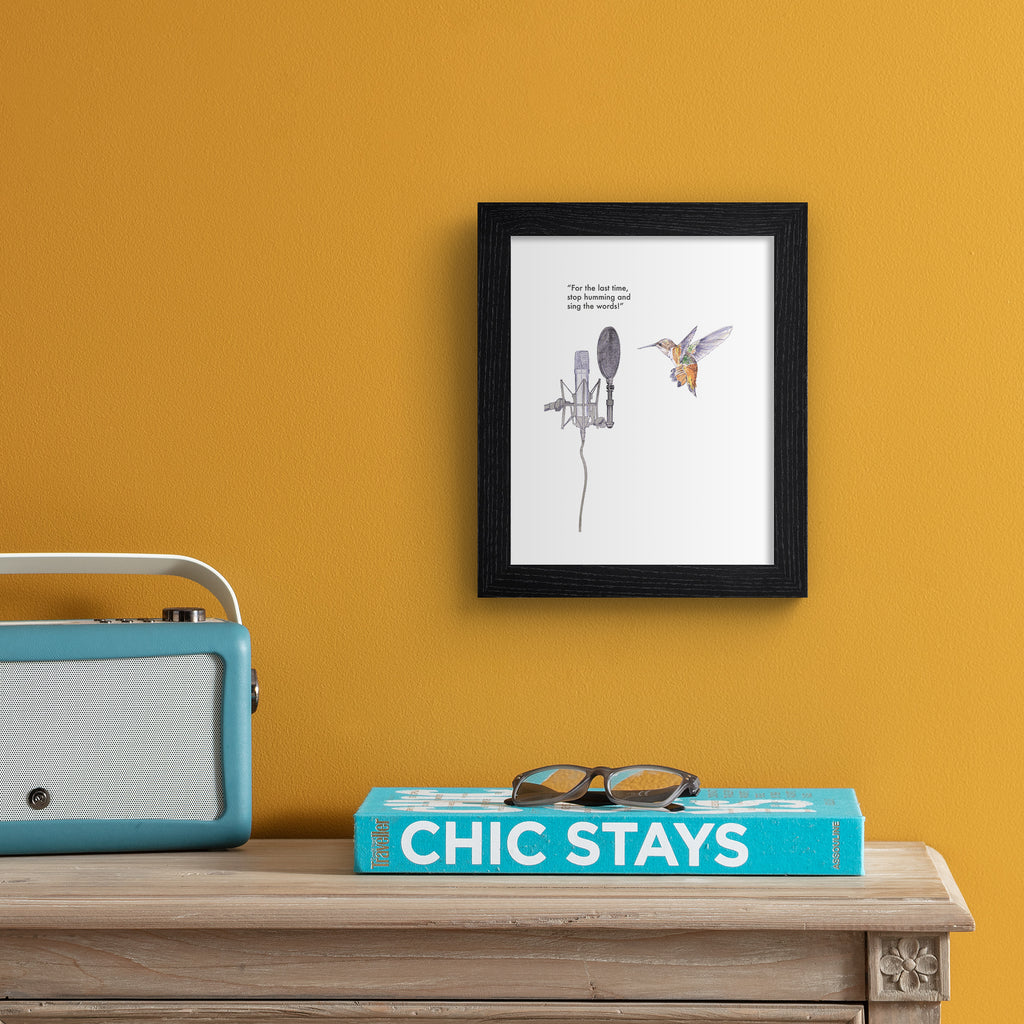 Humorous art print featuring an illustration of a hummingbird in front of a microphone. Text line reads 'For The Last Time, Stop Humming And Sing The Words!'. Art print is hung up on an orange wall.