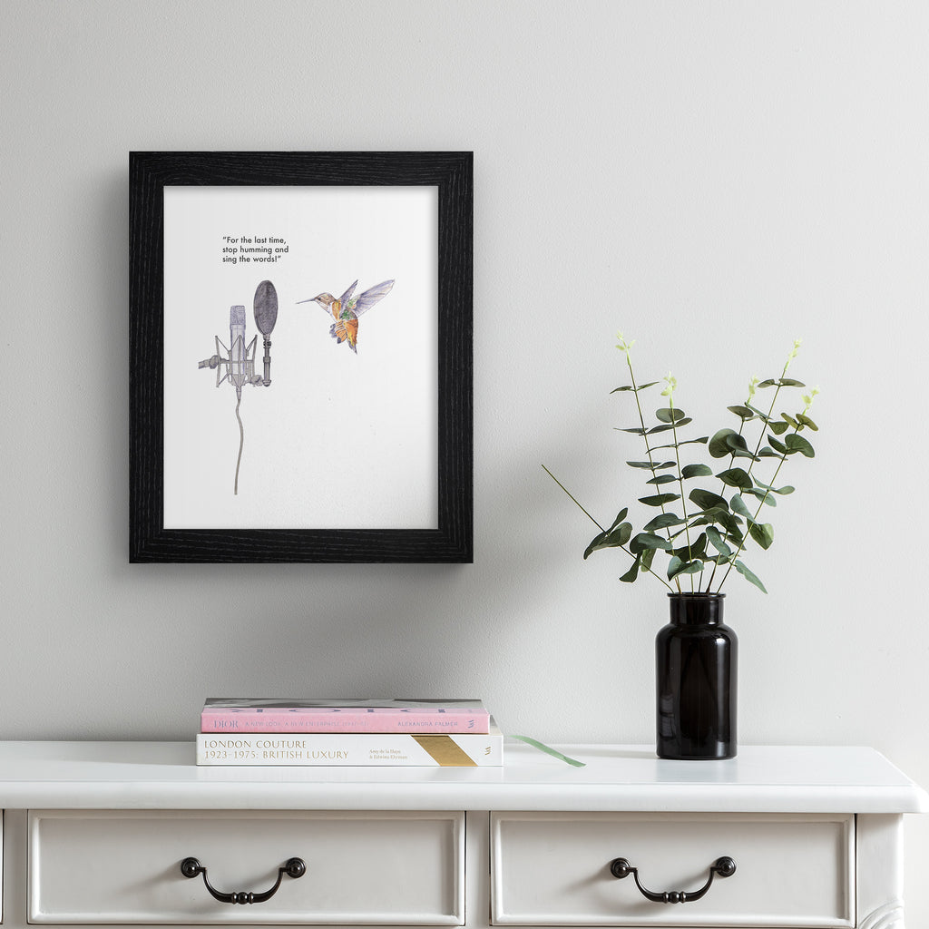 Humorous art print featuring an illustration of a hummingbird in front of a microphone. Text line reads 'For The Last Time, Stop Humming And Sing The Words!'. Art print is hung up on a pale grey wall.