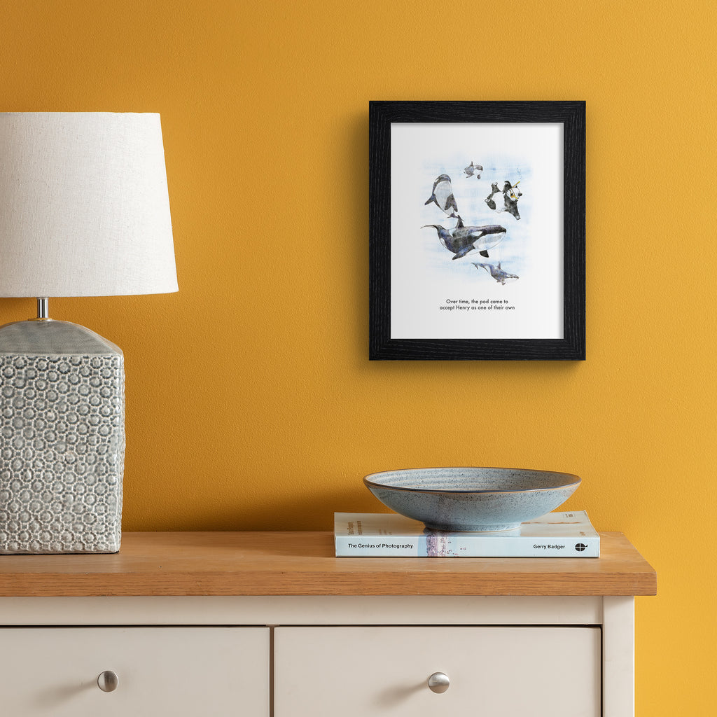 Humorous art print featuring an illustration of a panda swimming and 'blending' in with killer whales. Title underneath reads 'Over time, the pod came to accept Henry as one of their own'. Art print is hung up on an orange wall.