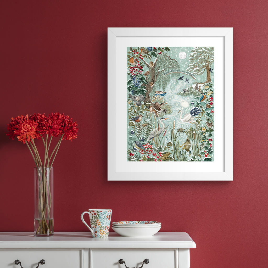 Christmas art print featuring a wintery wonderland scene of animals and botanicals basking by a river on a frosty day. Art print is hung up on a red wall.