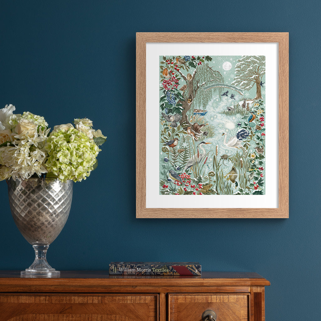 Christmas art print featuring a wintery wonderland scene of animals and botanicals basking by a river on a frosty day. Art print is hung up on a vibrant blue wall.