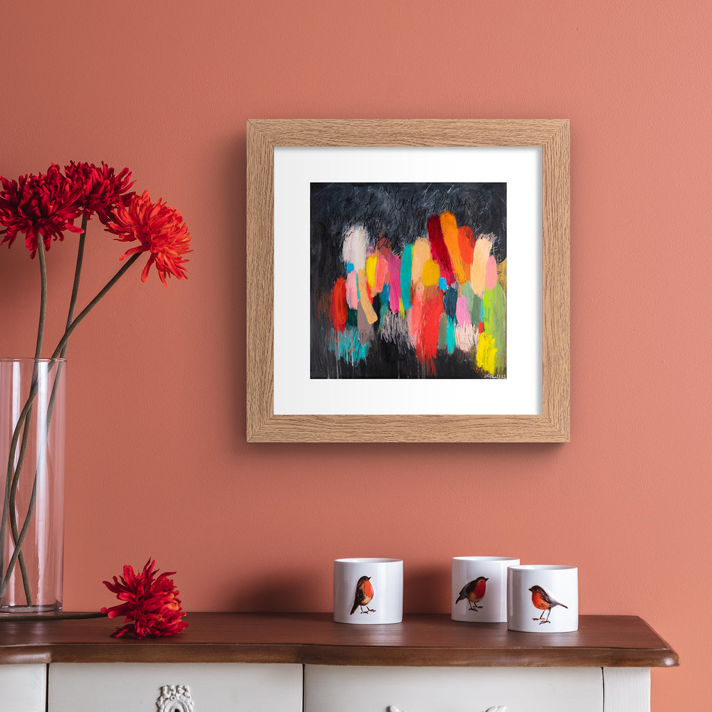 Vivid abstract print featuring a vast array of colours blended together on a moody background. Art print is hung up on a pink wall.