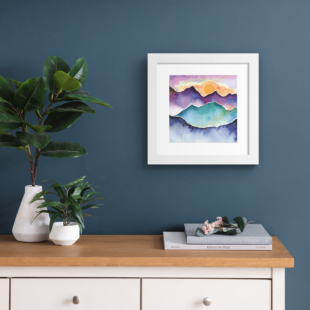 Watercolour art print featuring the sun rising above a beautiful mountain landscape. Art print is hung up on a dark blue wall.