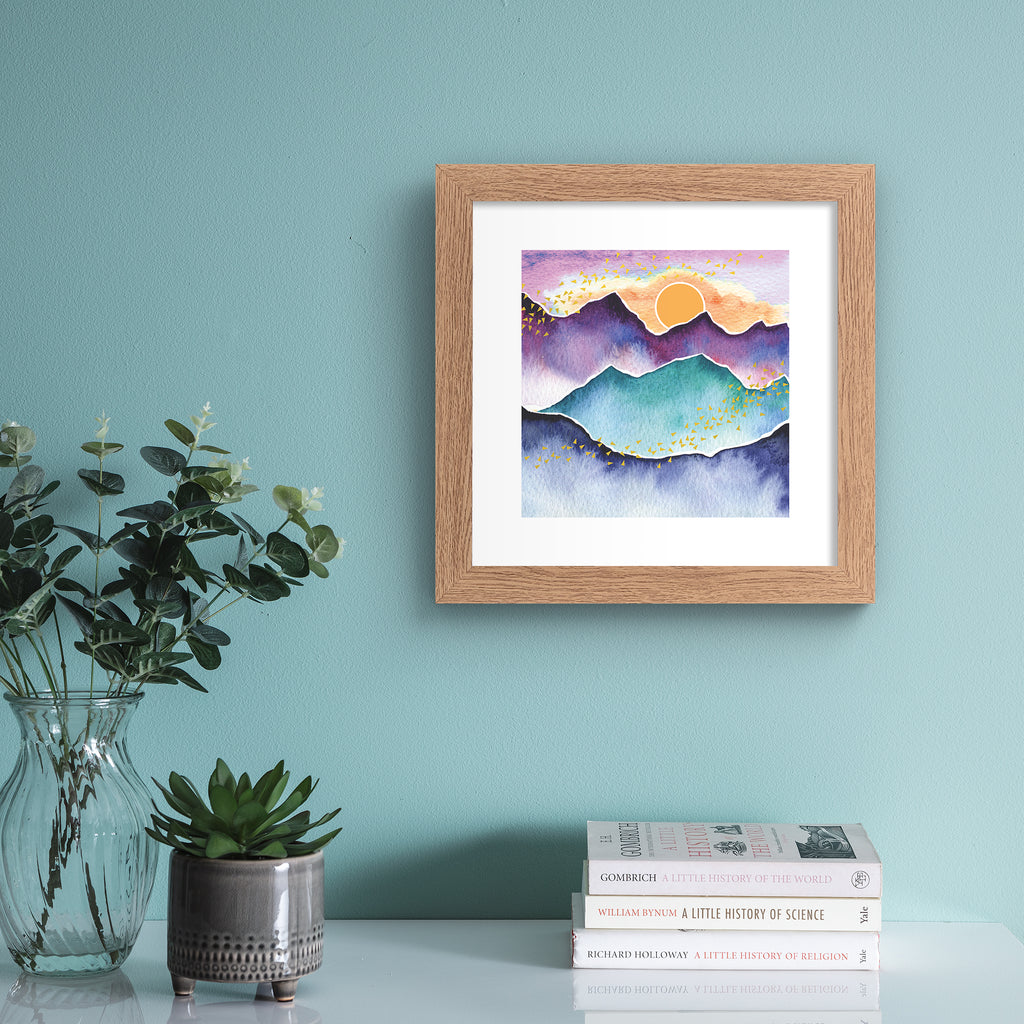 Watercolour art print featuring the sun rising above a beautiful mountain landscape. Art print is hung up on a light blue wall.