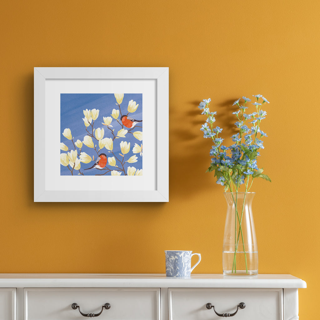 Stunning nature art print featuring two bullfinches perched in a blooming tree against a brilliant blue sky. Art print is hung up on an orange wall.
