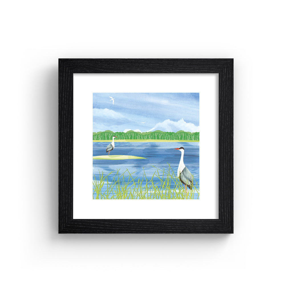 Nature art print featuring two herons standing on the edge of a bright blue lake, boarding a forest. Art print is in a black frame.
