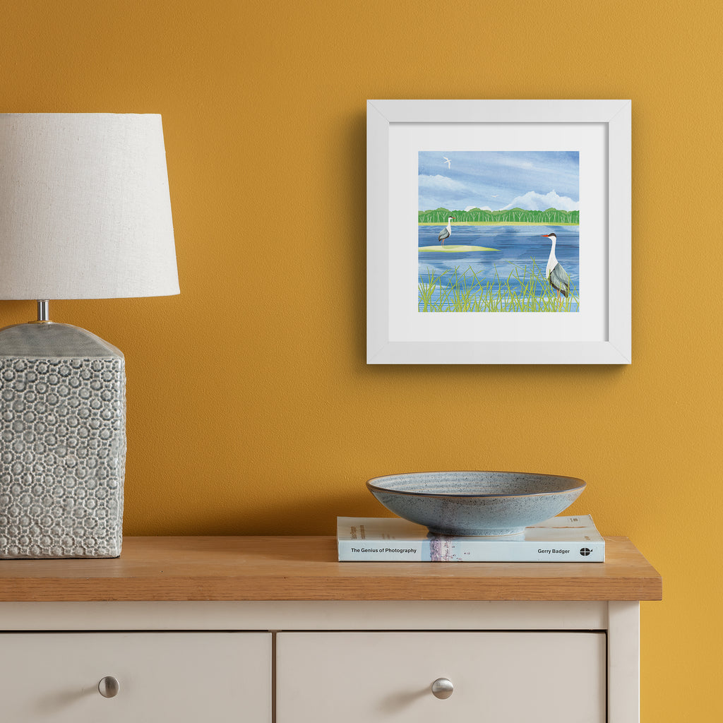 Nature art print featuring two herons standing on the edge of a bright blue lake, boarding a forest. Art print is hung up on an orange wall.