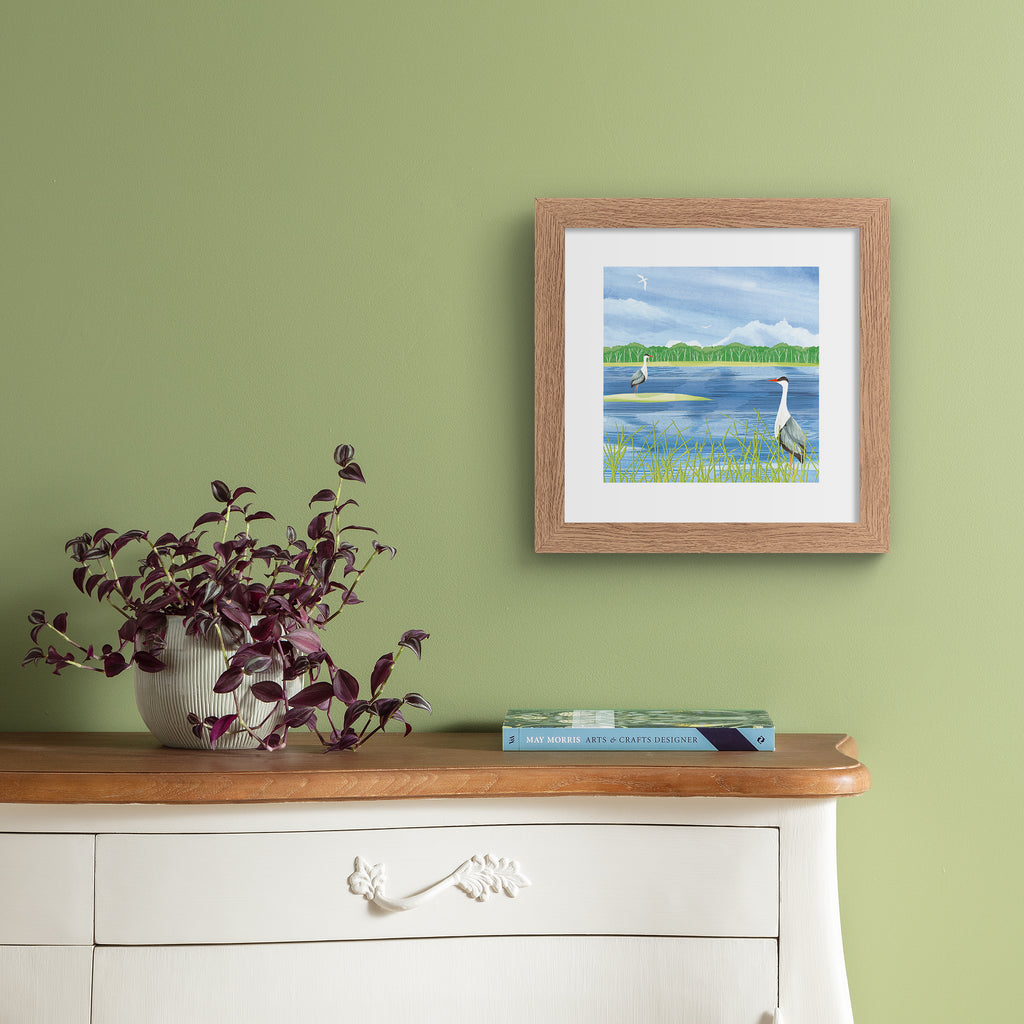 Nature art print featuring two herons standing on the edge of a bright blue lake, boarding a forest. Art print is hung up on a green wall.