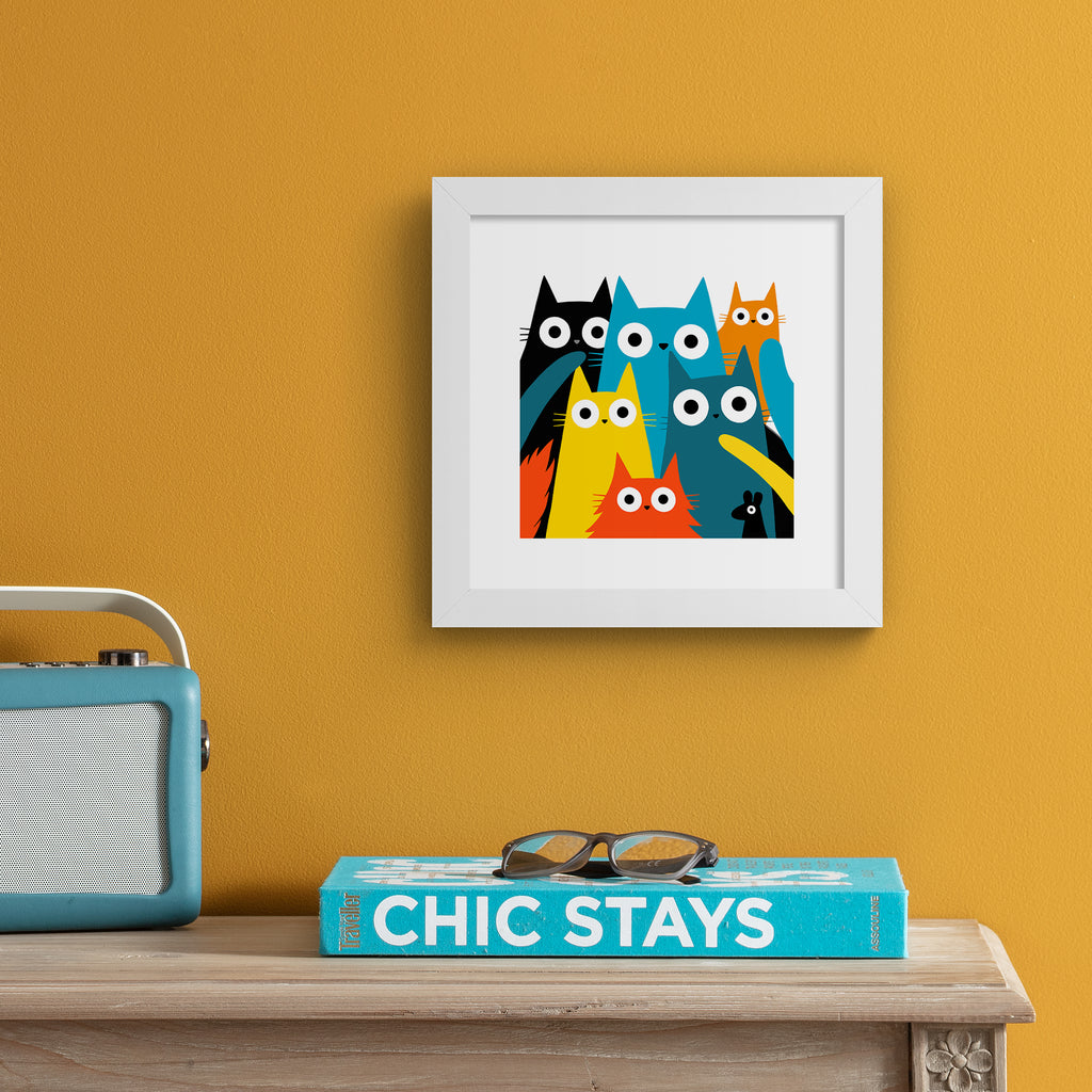 Colourful art print featuring different coloured cats and a mouse posing in the frame. Art print is hung up on an orange wall.