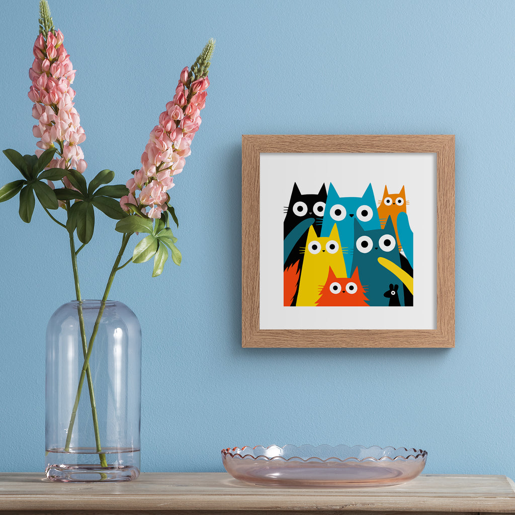 Colourful art print featuring different coloured cats and a mouse posing in the frame. Art print is hung up on a blue wall.