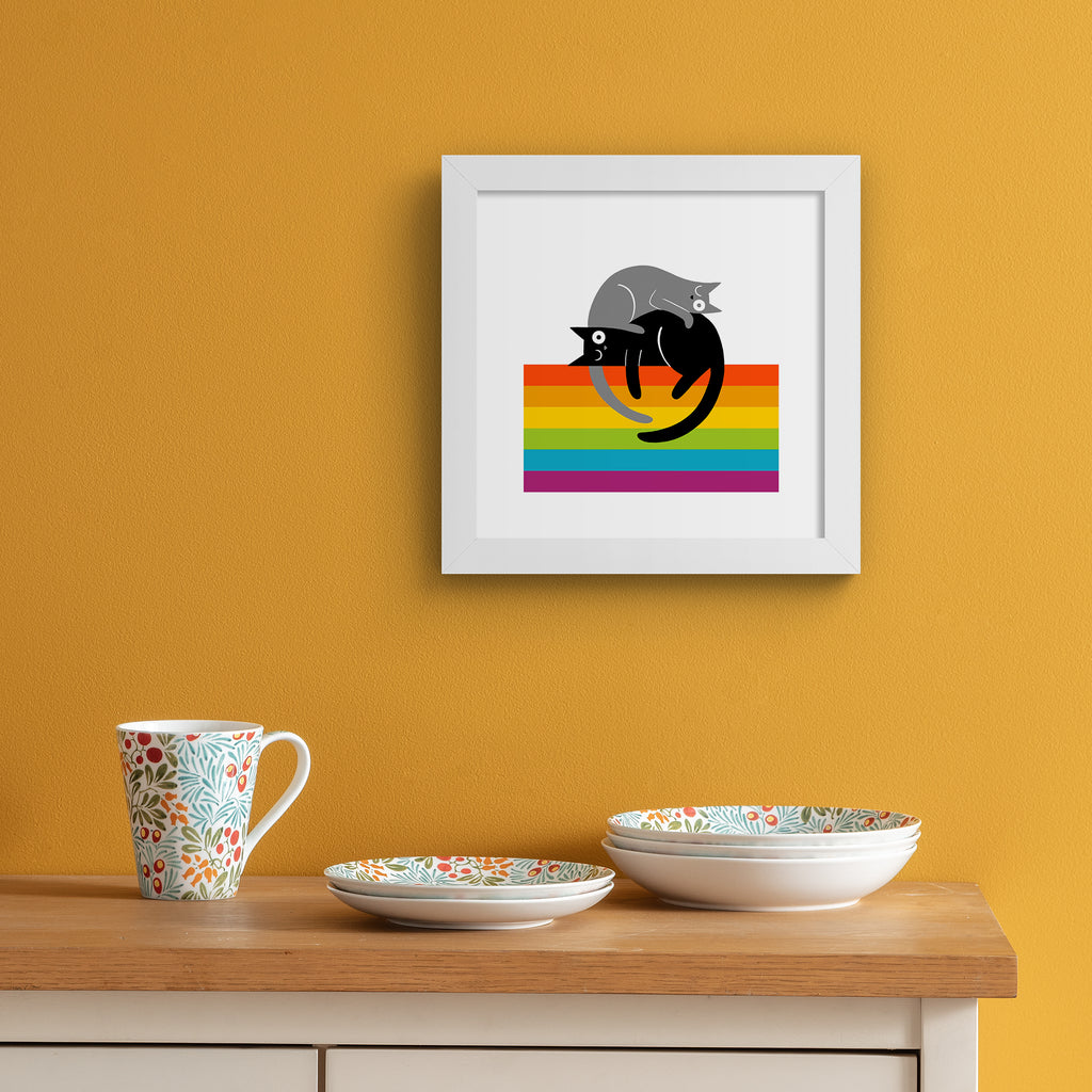 Art print of two kitties lying on top of a rainbow colours. Art print is hung up on an orange wall.