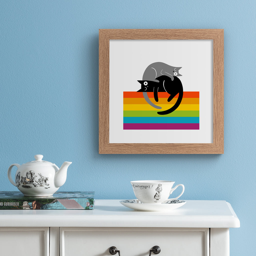 Art print of two kitties lying on top of a rainbow colours. Art print is hung up on a blue wall.